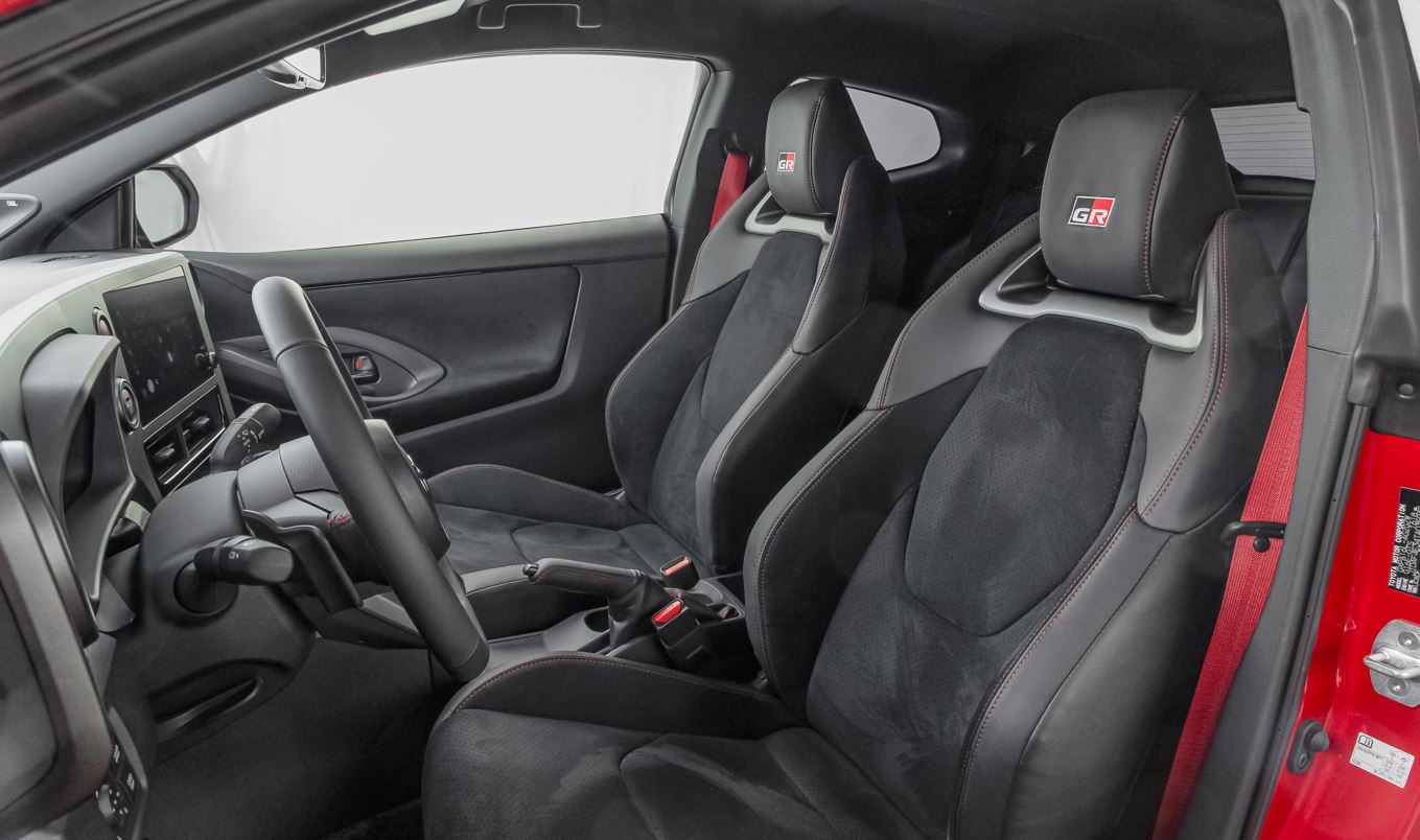 Interior do Toyota GR Yaris com bancos esportivos em couro e alcântara, cintos vermelhos e cockpit focado no motorista.