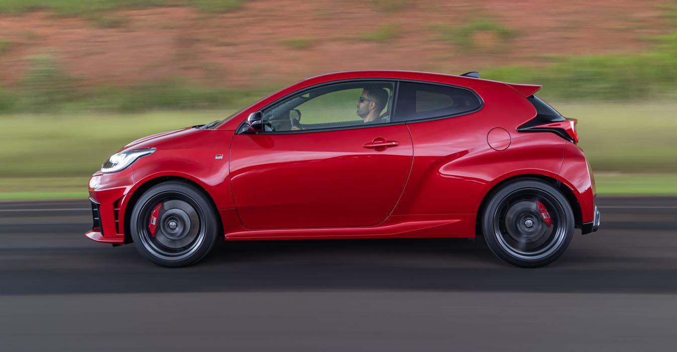 Toyota GR Yaris vermelho em perfil lateral, em movimento, destacando linhas musculosas, teto baixo e rodas esportivas em estrada.