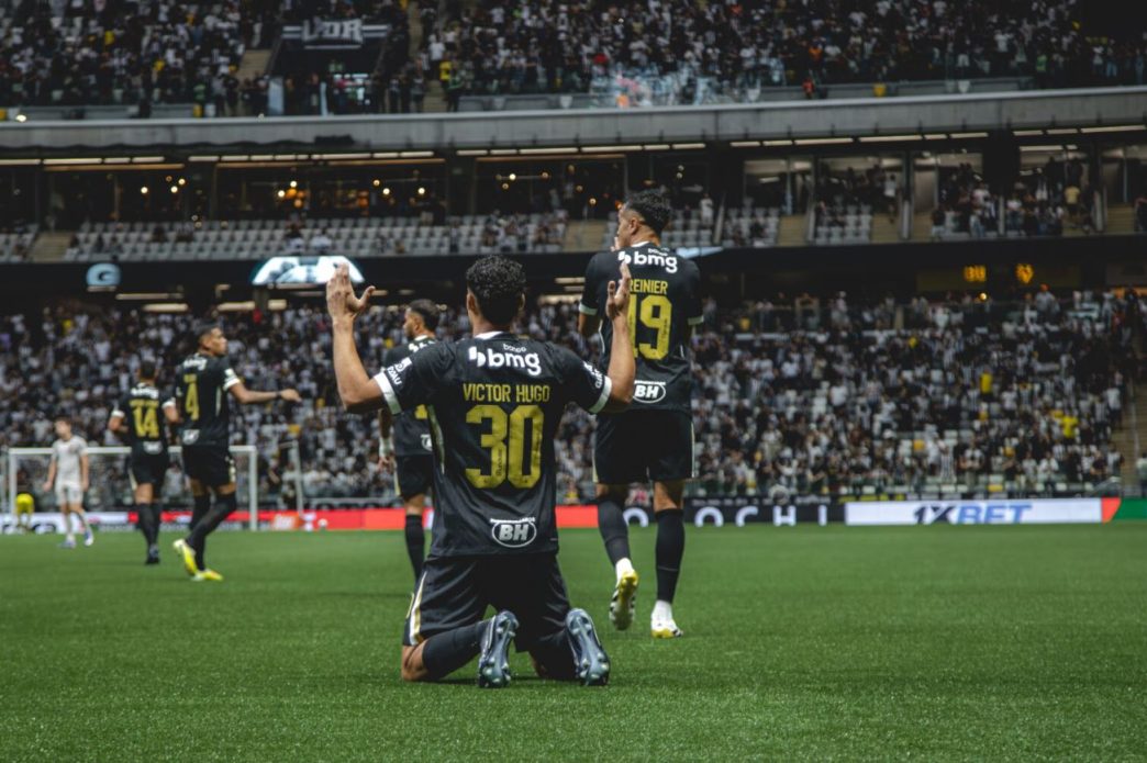 Jogador do Atlétioco, Victor Hugo, comemora gol ajoelhado no gramado da Arena MRV, com braços erguidos, enquanto companheiros celebram ao fundo diante da torcida.