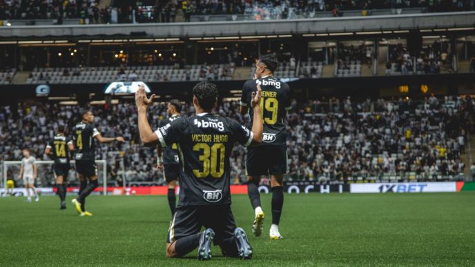 Jogador do Atlétioco, Victor Hugo, comemora gol ajoelhado no gramado da Arena MRV, com braços erguidos, enquanto companheiros celebram ao fundo diante da torcida.