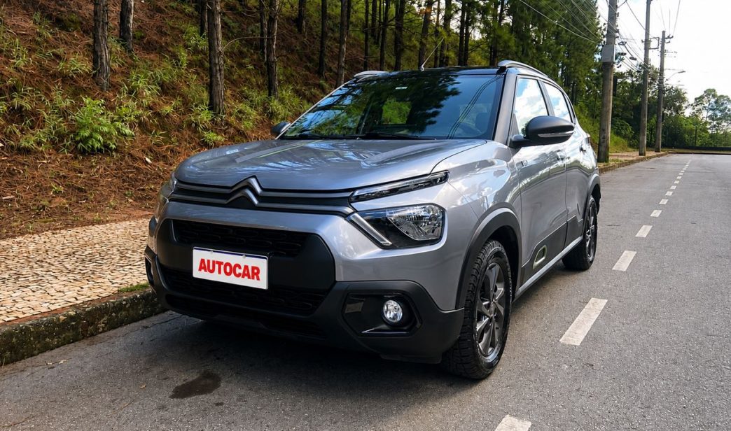 Vista dianteira em três quartos do Citroën C3 XTR 1.0 2026 na cor cinza, estacionado em via arborizada, destacando o visual aventureiro, rodas escurecidas e placa personalizada com a logomarca AUTOCAR.