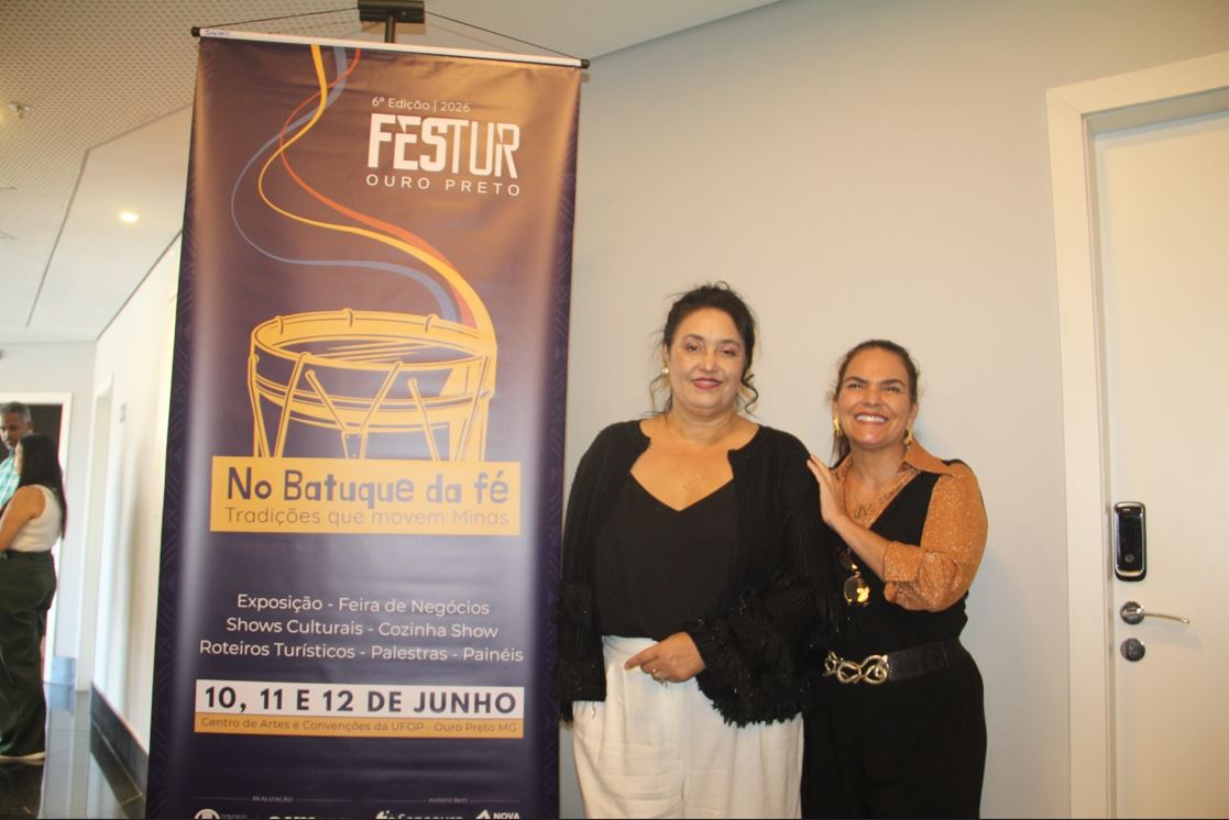 Duas mulheres posam sorrindo ao lado de um banner do FESTUR Ouro Preto 2026, com destaque para o tema “No Batuque da Fé – tradições que movem Minas” e informações do evento, em ambiente interno durante o lançamento.