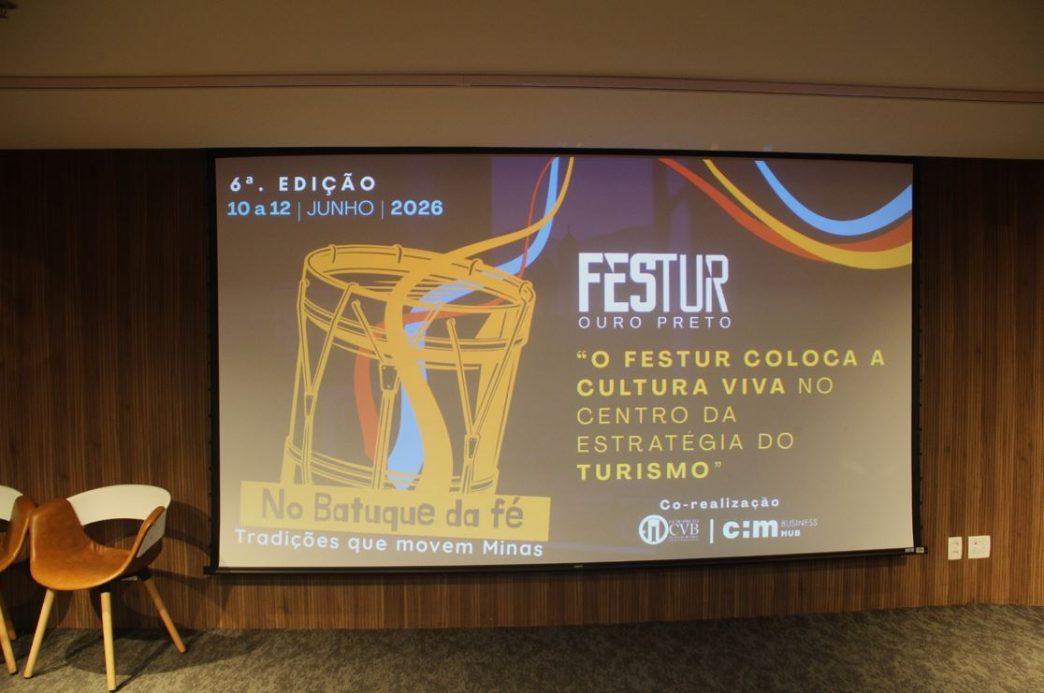 Tela de projeção em auditório exibe a identidade visual do FESTUR Ouro Preto 2026, com ilustração de tambor, datas do evento (10 a 12 de junho de 2026) e o tema “No Batuque da Fé – tradições que movem Minas”, ao lado de uma cadeira em ambiente institucional.