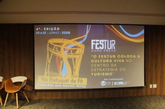 Tela de projeção em auditório exibe a identidade visual do FESTUR Ouro Preto 2026, com ilustração de tambor, datas do evento (10 a 12 de junho de 2026) e o tema “No Batuque da Fé – tradições que movem Minas”, ao lado de uma cadeira em ambiente institucional.
