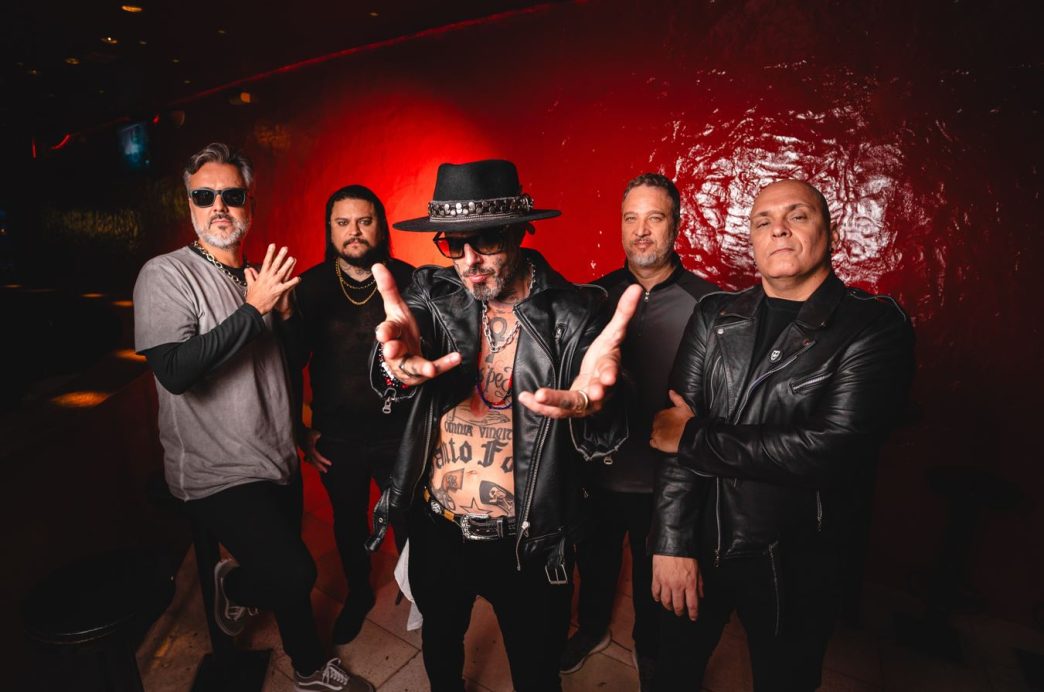 Integrantes da banda Detonautas posam em ambiente escuro, com parede vermelha ao fundo, em foto de divulgação do show em Belo Horizonte.