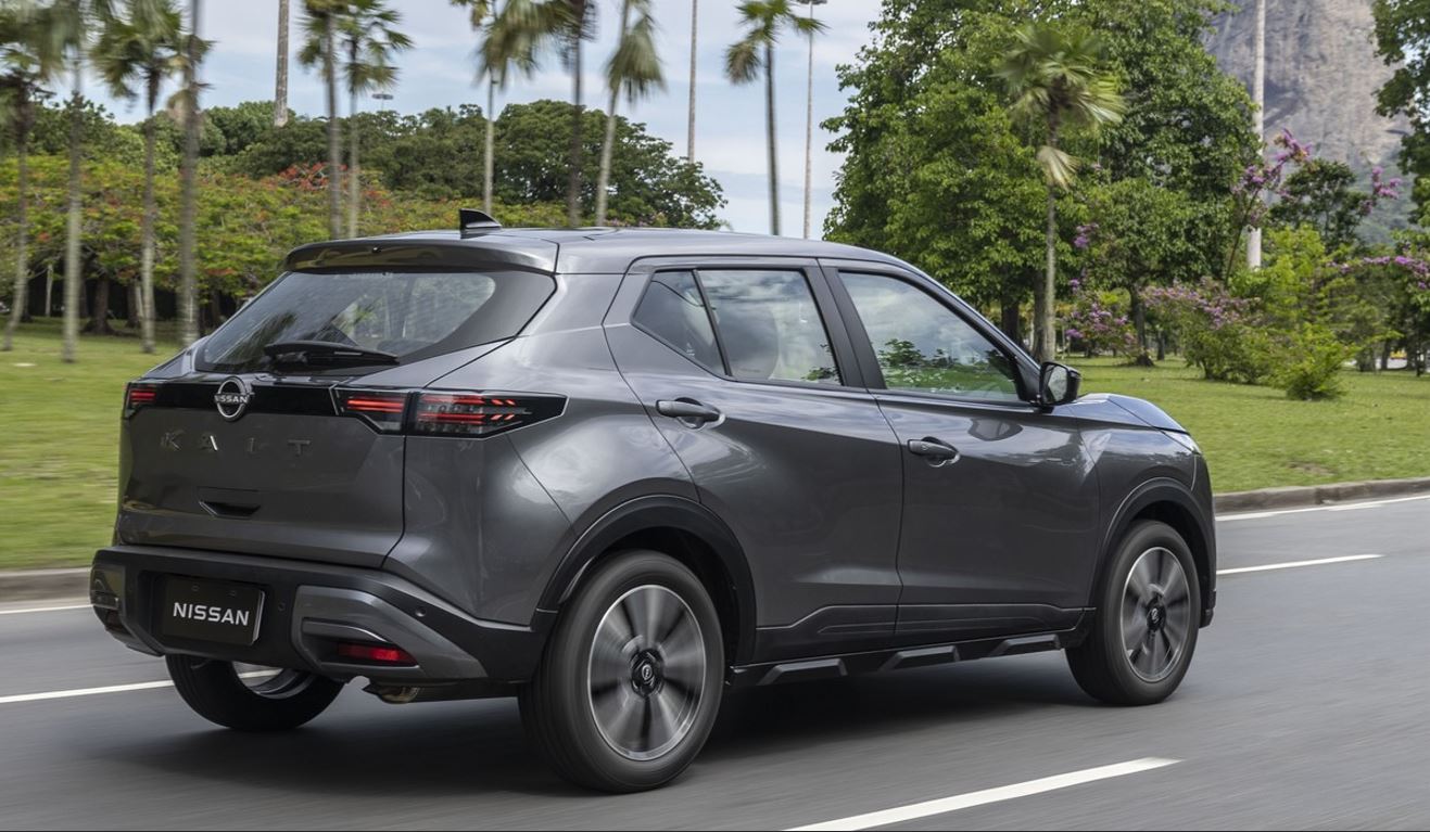 Nissan Kait cinza visto pela traseira em movimento, com lanternas em LED, nome do modelo na tampa do porta-malas, rodas de liga leve e carroceria de SUV compacto em avenida arborizada.