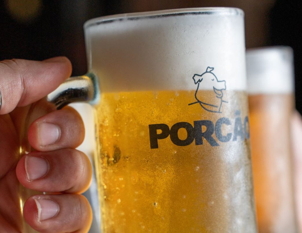 Caneca de chope gelado do Porcão BH em primeiro plano, segurada por uma pessoa, com outra bebida desfocada ao fundo.