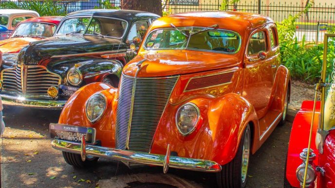 Carros antigos expostos em área arborizada, com destaque para um veículo clássico laranja em primeiro plano e outro preto ao fundo.