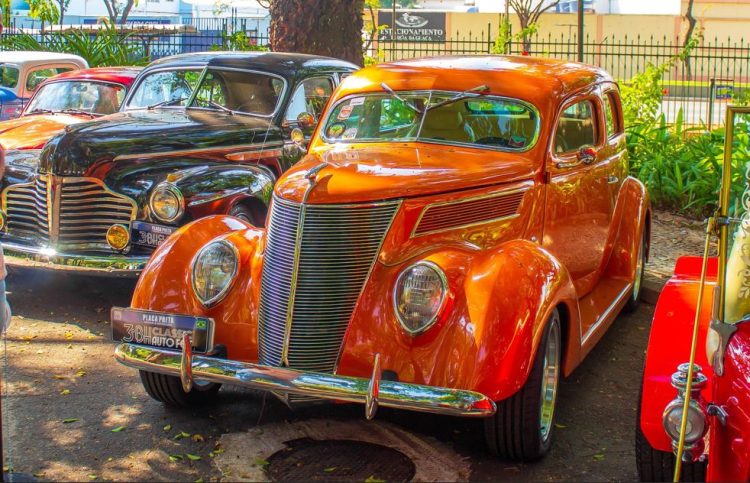 Carros antigos expostos em área arborizada, com destaque para um veículo clássico laranja em primeiro plano e outro preto ao fundo.