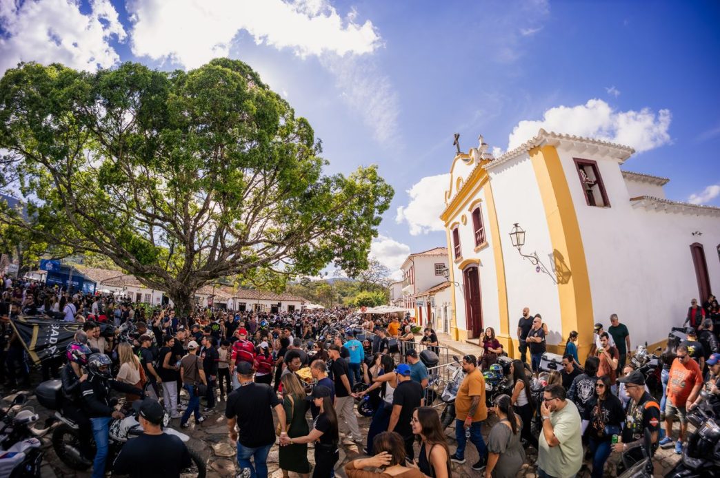 Multidão participa do Bike Fest Tiradentes no centro histórico da cidade, com motocicletas, visitantes e igreja colonial ao fundo.