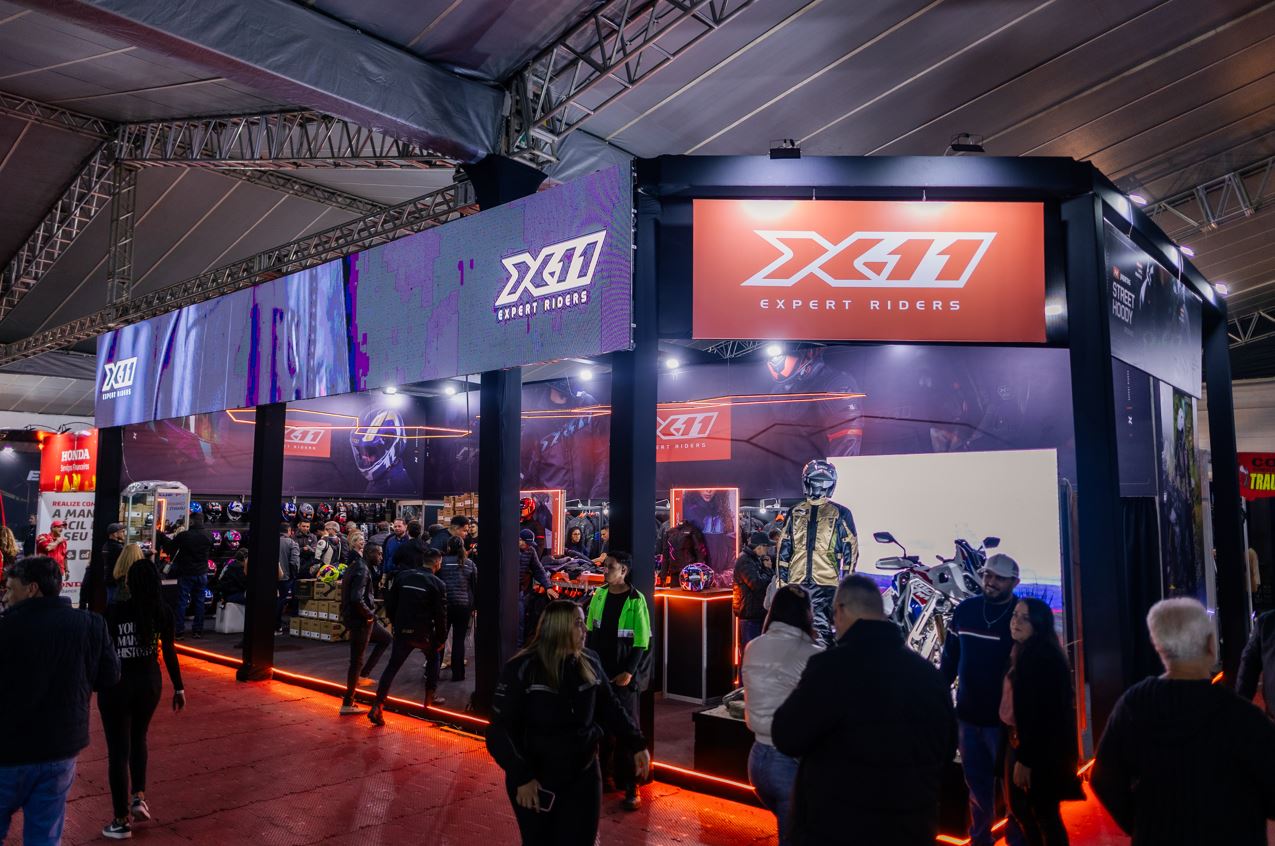 Visitantes circulam pelo estande da X11 durante a feira de negócios do Bike Fest Tiradentes, com produtos, motocicleta em exposição e painéis iluminados.