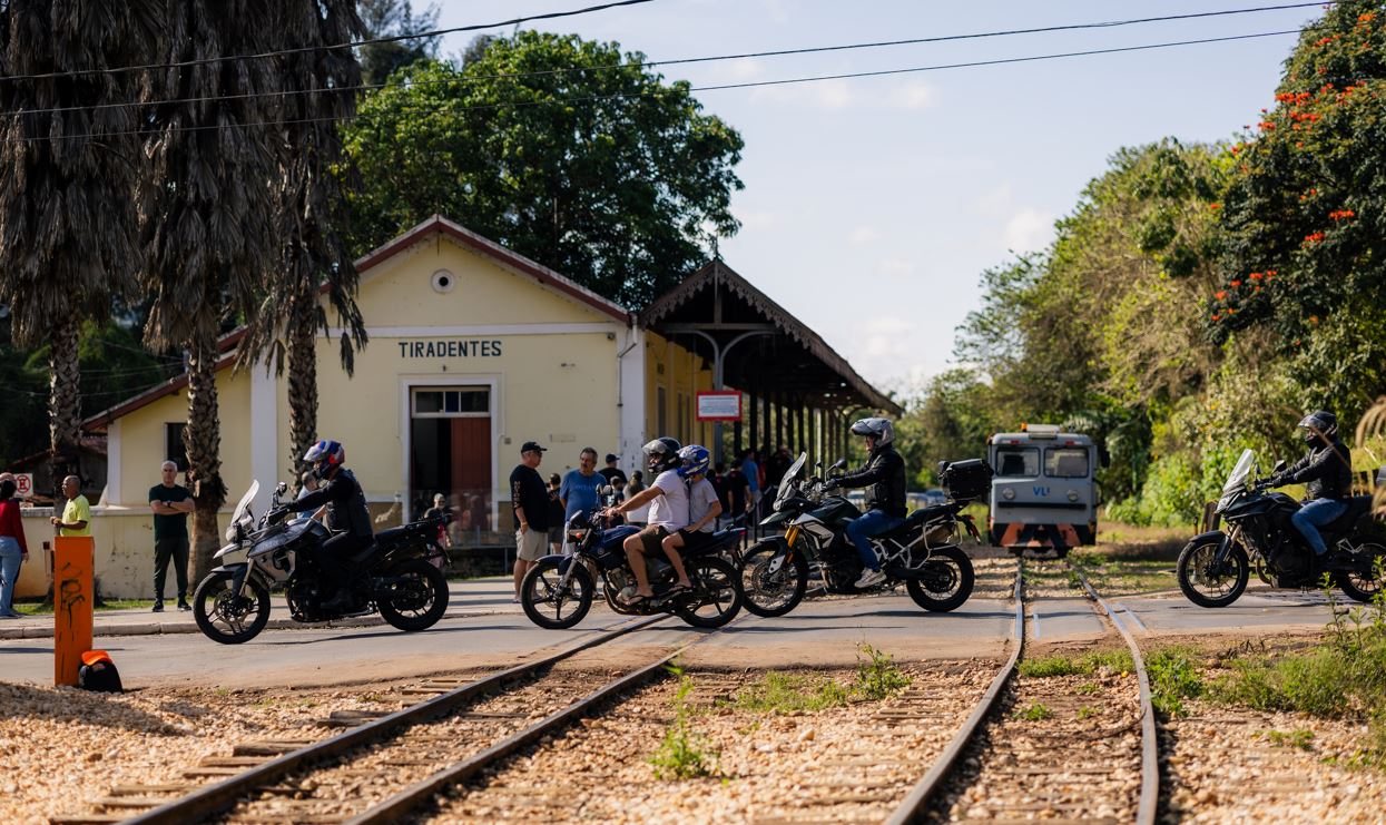 Motociclistas passam sobre trilhos em frente à estação ferroviária de Tiradentes durante o Bike Fest, com trem ao fundo e vegetação ao redor.