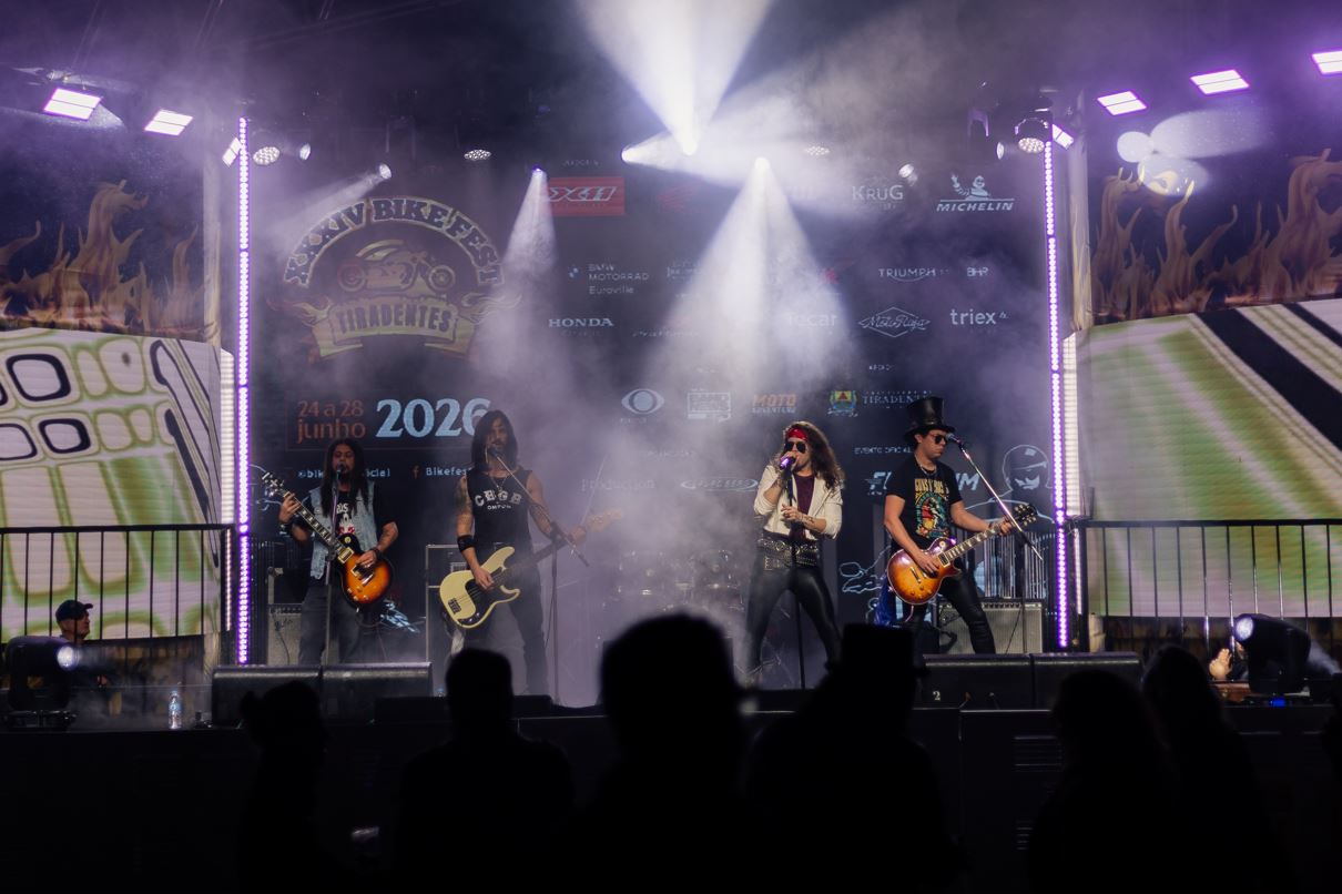 Banda de rock se apresenta no palco do Bike Fest Tiradentes, com iluminação intensa, fumaça cênica e público acompanhando o show. 