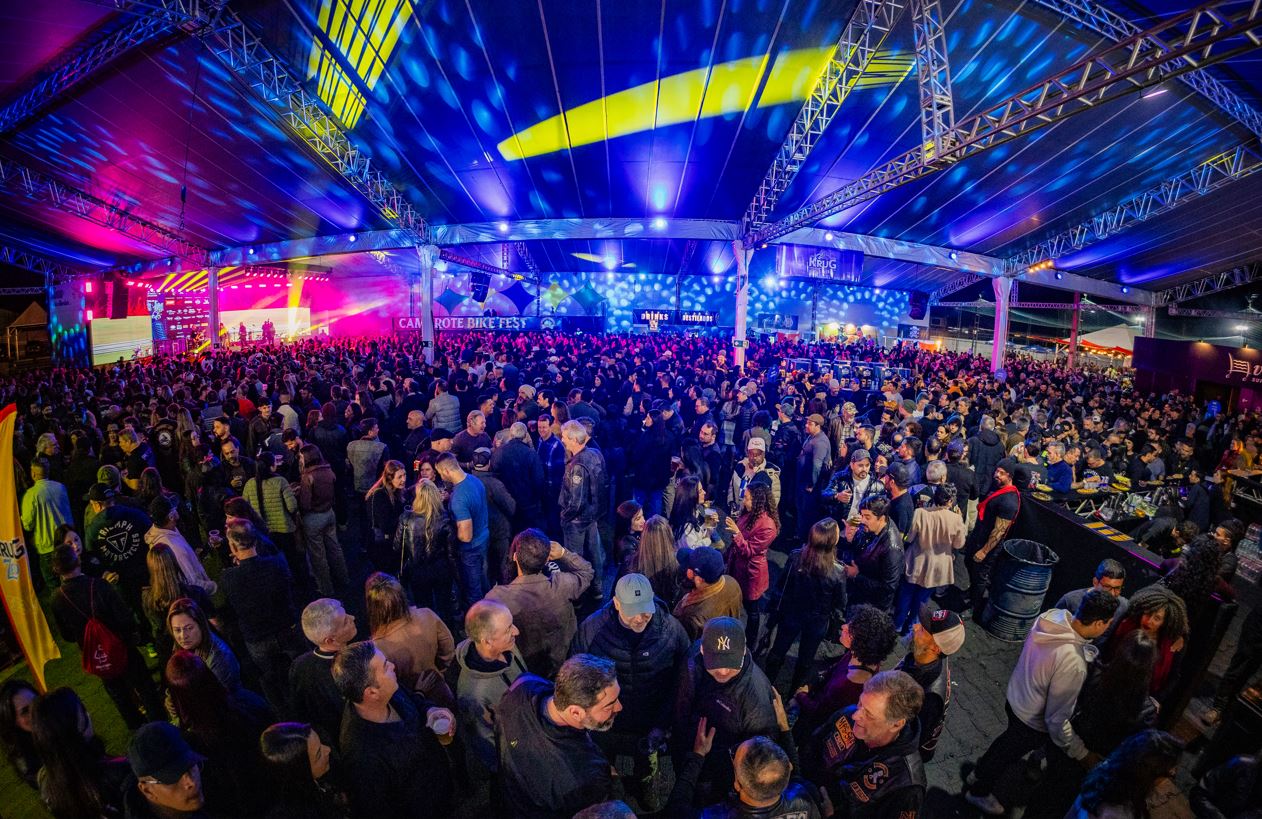 Público ocupa a área gastronômica do Bike Fest Tiradentes à noite, em espaço coberto com bares, iluminação colorida e palco ao fundo.