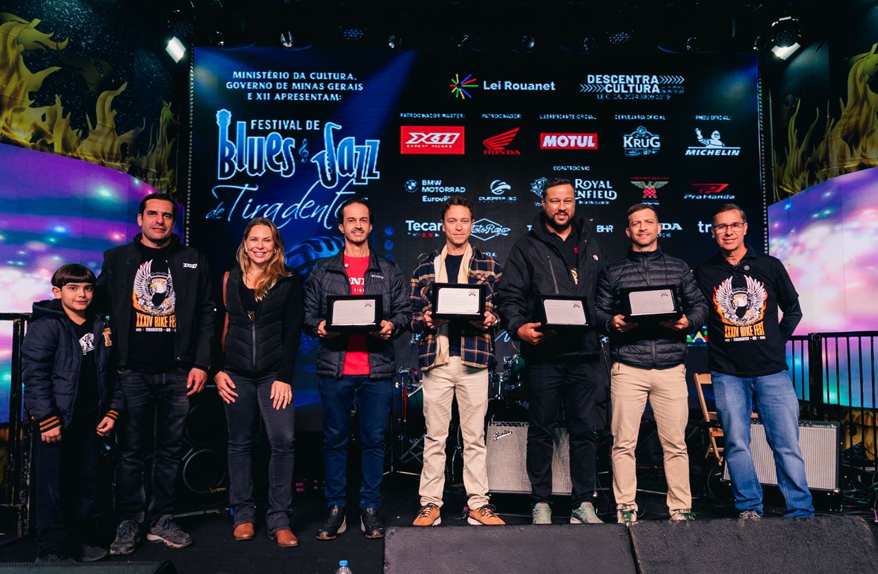 Homenageados posam no palco do Bike Fest Tiradentes segurando placas de reconhecimento, diante de painel com marcas apoiadoras do evento.