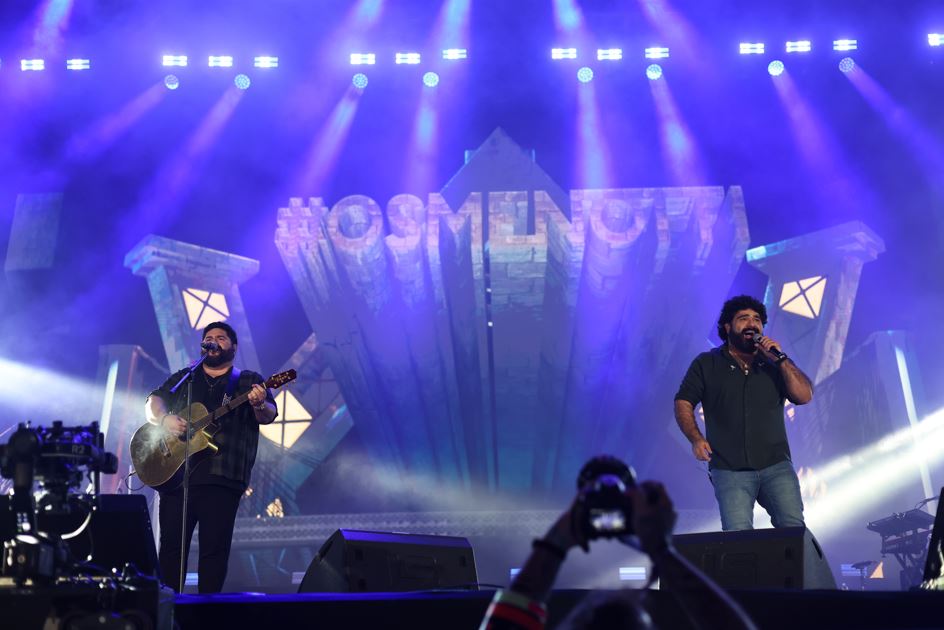 César Menotti e Fabiano se apresentam no palco do Fiat Festival 50 anos, com iluminação azul e público registrando o show.