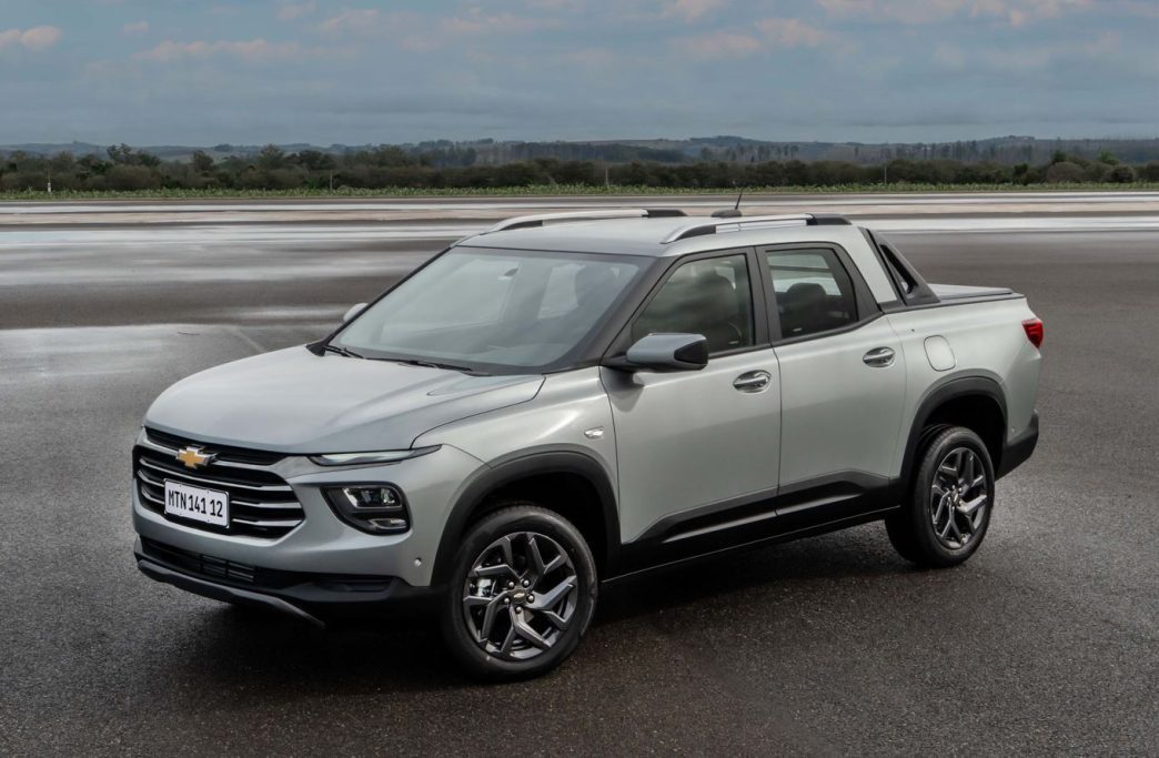 Chevrolet Montana 2027 prata em pista molhada, vista de frente e lateral, destacando visual de SUV com caçamba, rodas de liga leve, rack de teto e Santo Antônio integrado.