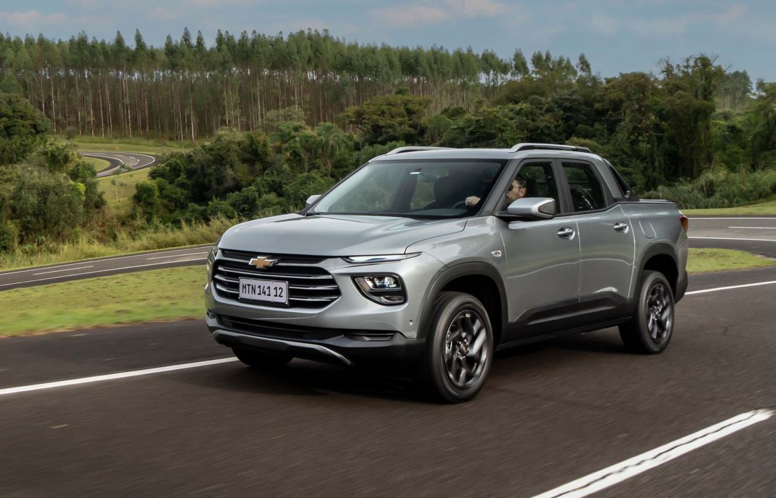 Chevrolet Montana 2027 cinza em movimento na estrada, vista de frente e lateral, destacando grade frontal, faróis em LED, rodas de liga leve e perfil de SUV com caçamba.
