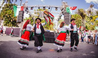 Dançarinos com trajes típicos italianos se apresentam diante de palco decorado com bandeiras da Itália.