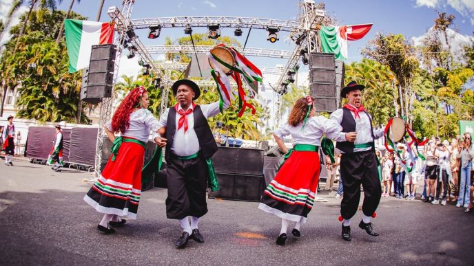 Dançarinos com trajes típicos italianos se apresentam diante de palco decorado com bandeiras da Itália.