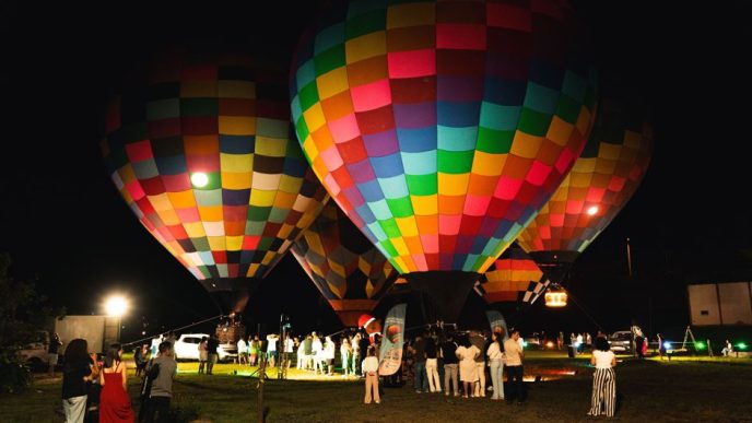 Balões coloridos iluminados à noite permanecem próximos ao solo enquanto visitantes acompanham a atração em uma área aberta.