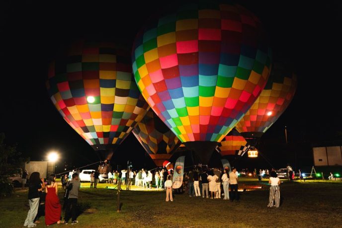 Balões coloridos iluminados à noite permanecem próximos ao solo enquanto visitantes acompanham a atração em uma área aberta.