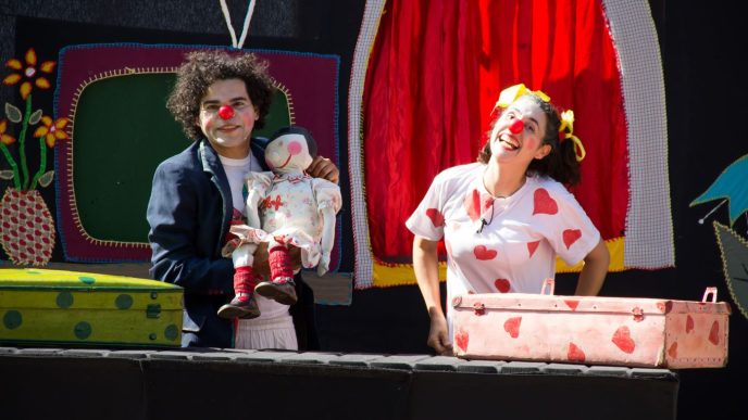 Dois artistas caracterizados como palhaços apresentam espetáculo teatral com boneco e cenário colorido em cena do grupo Yepocá.