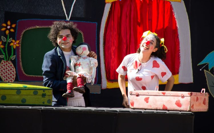 Dois artistas caracterizados como palhaços apresentam espetáculo teatral com boneco e cenário colorido em cena do grupo Yepocá.