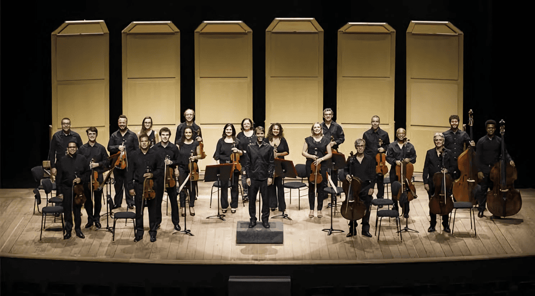 Integrantes da Orquestra SESIMINAS posam no palco com violinos, violas, violoncelos e contrabaixos, diante de painéis acústicos.