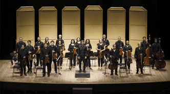 Integrantes da Orquestra SESIMINAS posam no palco com violinos, violas, violoncelos e contrabaixos, diante de painéis acústicos.