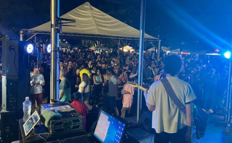 Vista do palco durante apresentação de rock, com músico de costas tocando guitarra diante de um grande público reunido em evento noturno.