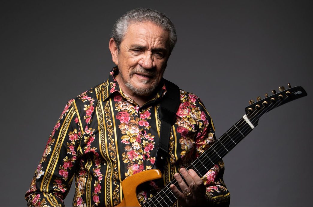 Zé Ramalho posa diante de um fundo escuro, segurando uma guitarra preta e usando uma camisa estampada com flores e detalhes dourados.