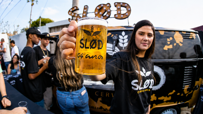 Mulher ergue caneca de chope da Slod durante evento, diante de veículo personalizado e público ao ar livre.