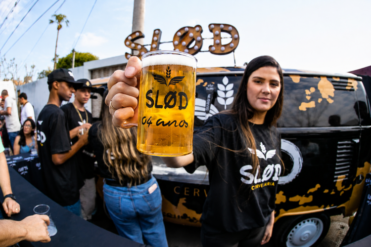 Mulher ergue caneca de chope da Slod durante evento, diante de veículo personalizado e público ao ar livre.