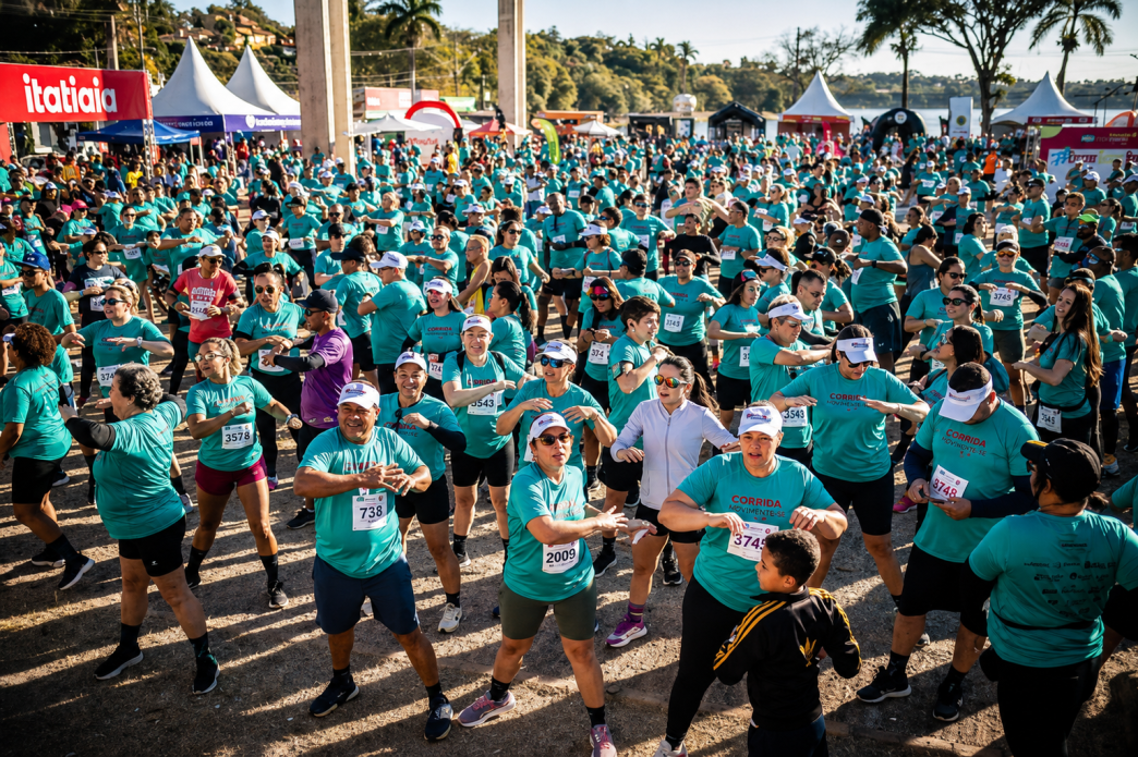Grande grupo de corredores com camisetas azul-turquesa participa de atividade coletiva em arena da Corrida Itatiaia, em Belo Horizonte, com estruturas do evento ao fundo.