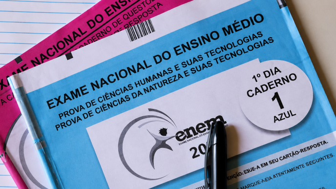 Cadernos de prova do ENEM nas cores azul e rosa sobre uma mesa, acompanhados de caneta preta e folha pautada.