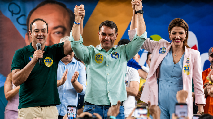 Flávio Roscoe, Flávio Bolsonaro e Ellen Miziara aparecem de mãos erguidas durante evento político, diante do público, em Belo Horizonte.