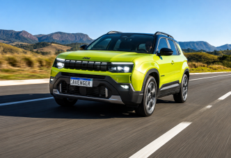 Jeep Avenger verde em movimento por uma rodovia, com montanhas ao fundo, destacando a grade de sete fendas, faróis em LED e rodas de liga leve.