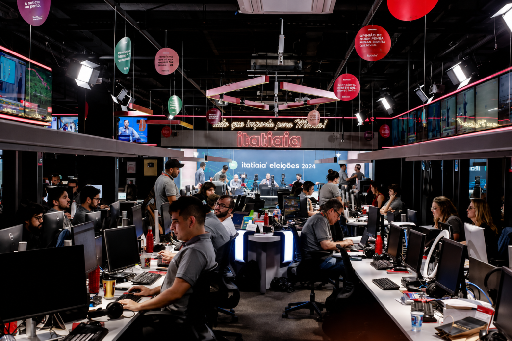 Redação da Rádio Itatiaia em Belo Horizonte, com jornalistas e profissionais trabalhando em computadores e estúdios integrados.