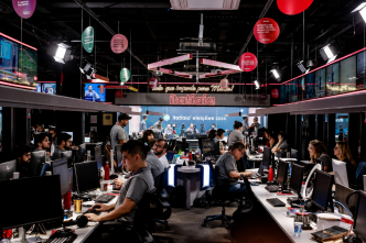 Redação da Rádio Itatiaia em Belo Horizonte, com jornalistas e profissionais trabalhando em computadores e estúdios integrados.