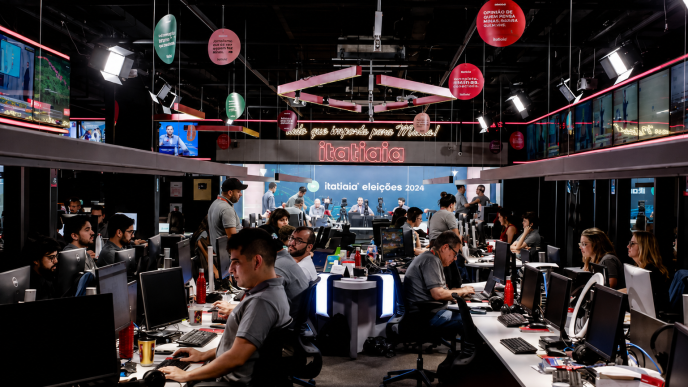 Redação da Rádio Itatiaia em Belo Horizonte, com jornalistas e profissionais trabalhando em computadores e estúdios integrados.