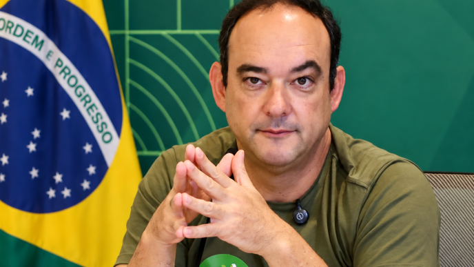 Flávio Roscoe sentado à mesa, diante da bandeira do Brasil, usando camiseta verde com adesivo de campanha “Flávio 22”.