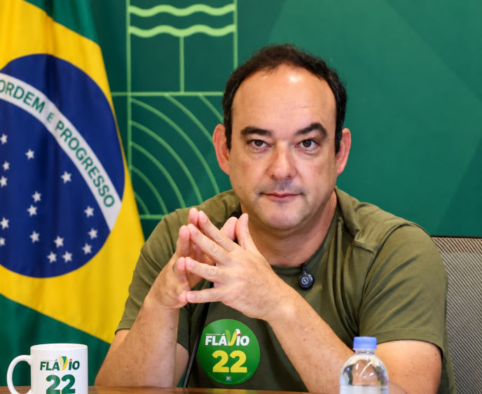 Flávio Roscoe sentado à mesa, diante da bandeira do Brasil, usando camiseta verde com adesivo de campanha “Flávio 22”.