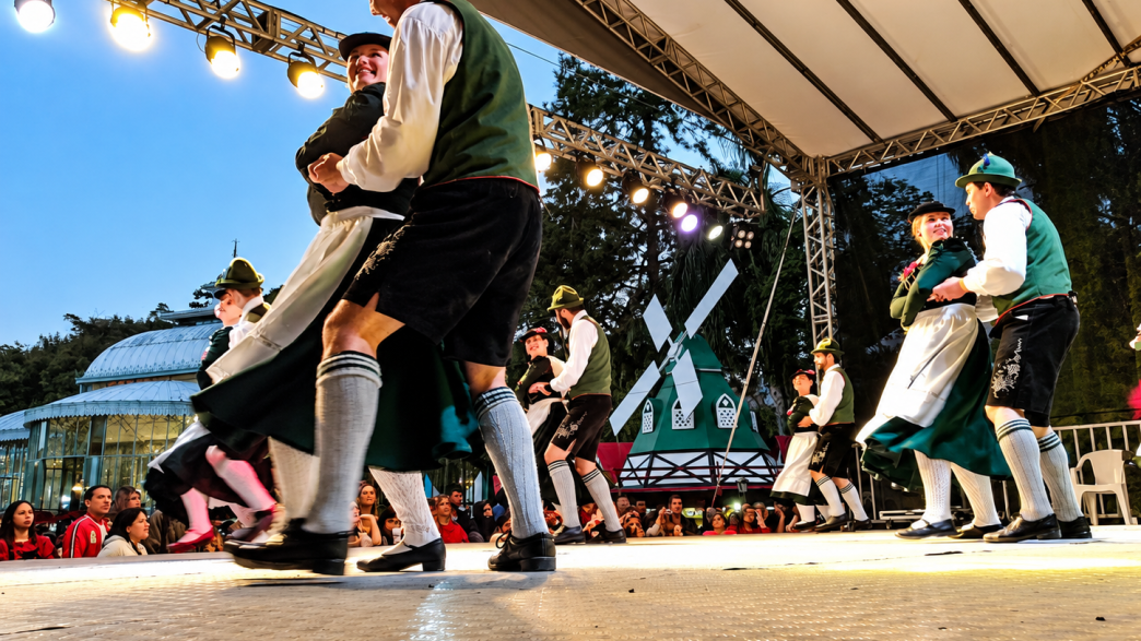 Casais com trajes típicos bávaros apresentam dança folclórica alemã em palco iluminado, representando as tradições culturais da Oktoberfest Betim 2026.