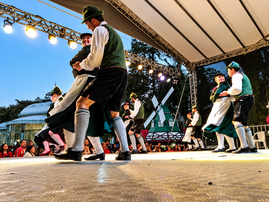 Casais com trajes típicos bávaros apresentam dança folclórica alemã em palco iluminado, representando as tradições culturais da Oktoberfest Betim 2026.