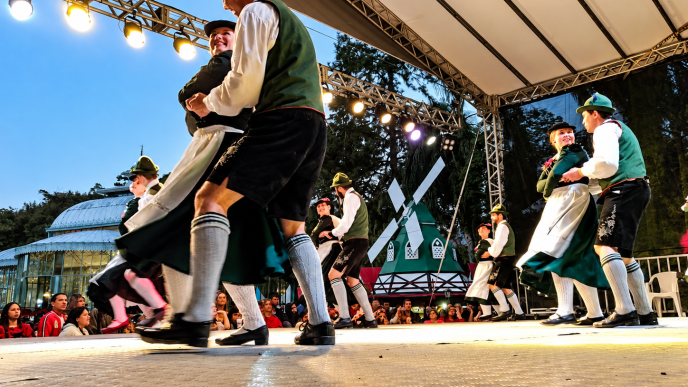 Casais com trajes típicos bávaros apresentam dança folclórica alemã em palco iluminado, representando as tradições culturais da Oktoberfest Betim 2026.