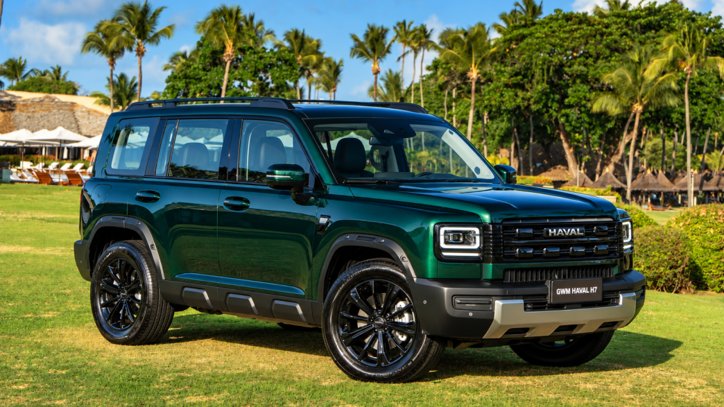 GWM Haval H7 híbrido plug-in flex na cor Verde Tijuca, visto de frente e lateral, com grade robusta, faróis de LED e rodas pretas, estacionado em resort tropical com palmeiras ao fundo.