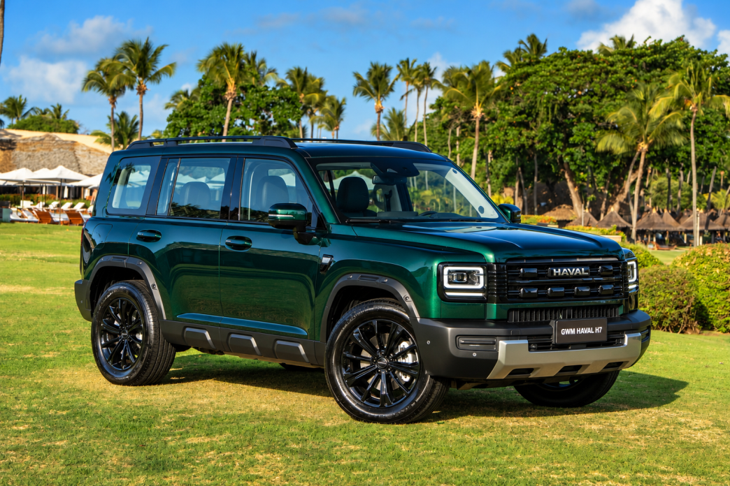 GWM Haval H7 híbrido plug-in flex na cor Verde Tijuca, visto de frente e lateral, com grade robusta, faróis de LED e rodas pretas, estacionado em resort tropical com palmeiras ao fundo.