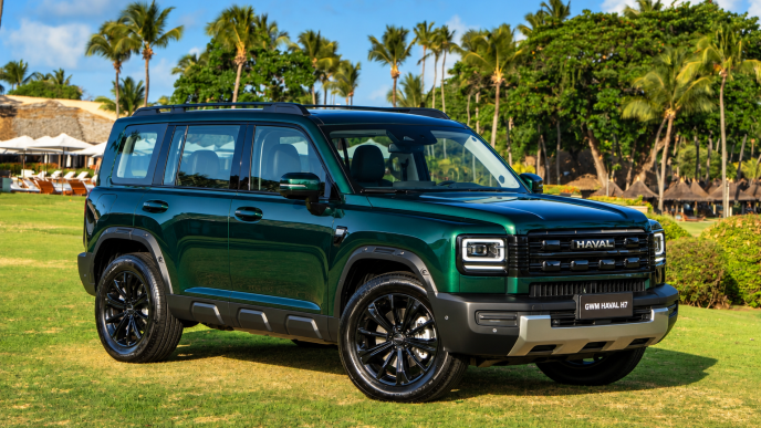 GWM Haval H7 híbrido plug-in flex na cor Verde Tijuca, visto de frente e lateral, com grade robusta, faróis de LED e rodas pretas, estacionado em resort tropical com palmeiras ao fundo.