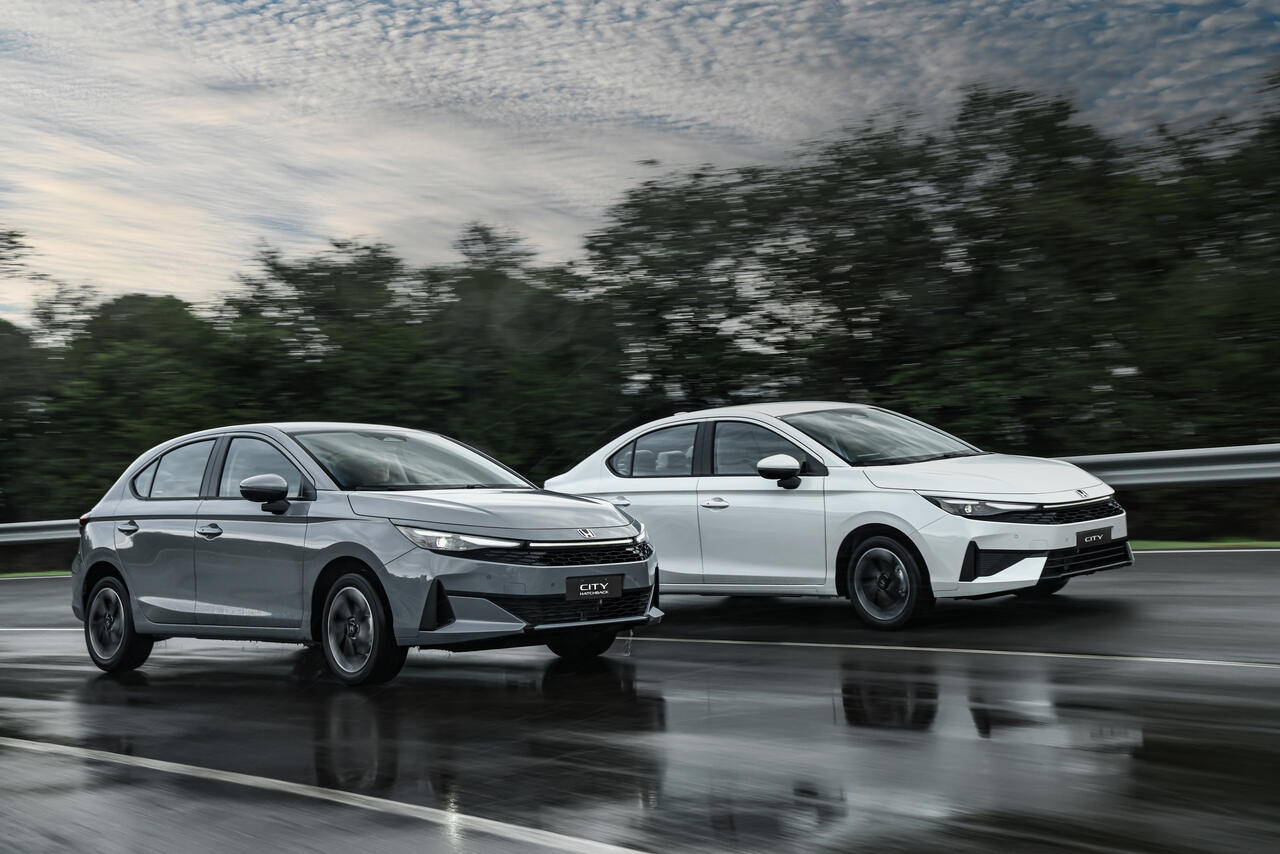 Honda City 2027 Hatchback cinza e Sedan branco em movimento na pista molhada, destacando nova dianteira, grade frontal e faróis Full LED.